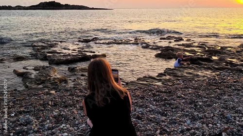 A woman on a pebble beach takes pictures of a child playing by the sea on her smartphone against the background of a warm sunset.