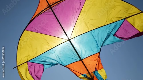 Vibrant Colorful Diamond Kite Soaring Against a Bright Sunny Blue Sky with Sun Peeking Through Fabric
