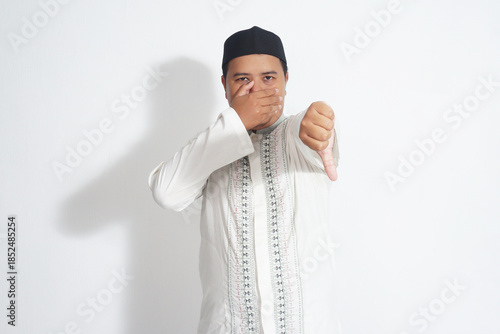 an Indonesian Muslim man wearing a black peci and a white koko shirt is covering his mouth while giving a thumbs down to a plain white background