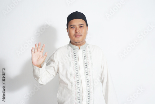 an Indonesian Muslim man wearing a black peci and white koko shirt is raising his hand while saying no on isolated white background