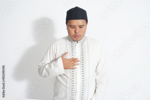 An Indonesian Muslim man wearing a black peci, white mask, and white koko shirt is stroking his chest to be patient on a plain white background.