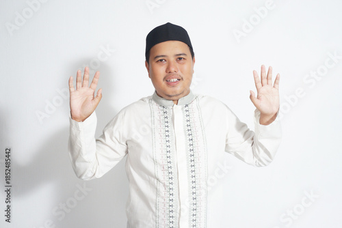 An Indonesian Muslim man wearing a black peci and white koko shirt posing is open the hand for saying the hi on a plain white background.                