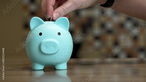 student deposits coin in piggy bank, blue piggy bank sits on kitchen counter, closeup of student placing coin into piggy bank, warm mosaic backsplash frames student saving small change patiently