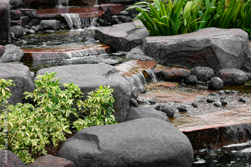 Fotografi A detailed view of a multi-tiered garden water feature, featuring water cascading over and around large, dark gray rocks