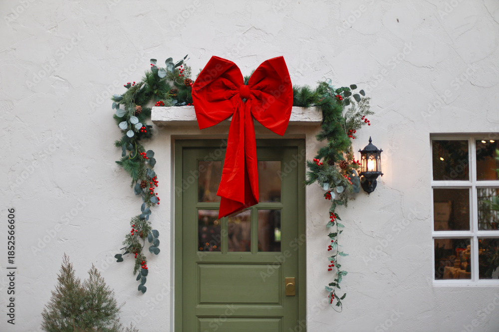Fototapeta premium Merry Christmas decoration with large red bow and green garland over front door of white house exterior creating festive and welcoming holiday atmosphere for winter celebration season