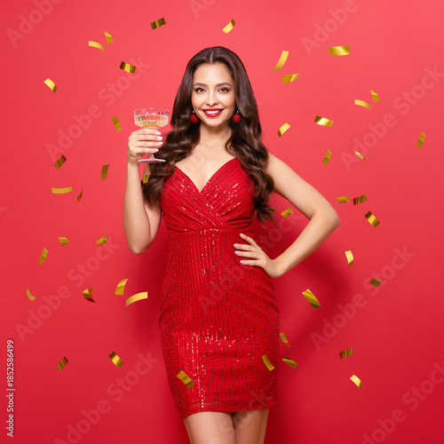 Beautiful woman in cocktail dress posing with glass of champagne under confetti. Model with curly hair. Christmas or Happy New Year. Holiday and Party