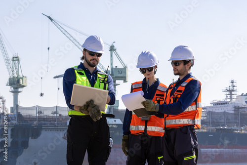 group of industrial engineers workers in a refinery - oil and gas processing equipment and machinery, engineers collaborate with a laptop, blueprint, and digital tablet at the oil storage tanks site.