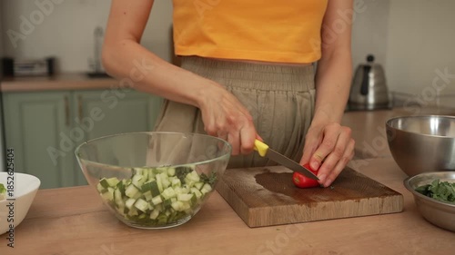 Close-up of female hands cutting fresh vegetables on a wooden board in a modern kitchen. The woman is chopping a ripe red tomato for a healthy, nutritious salad with sliced cucumbers
