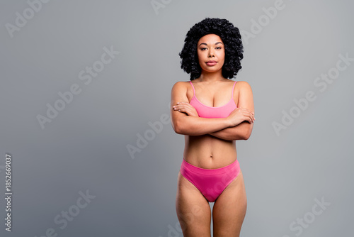 Bright confident young woman in pink bikini stands with arms crossed against a grey studio background