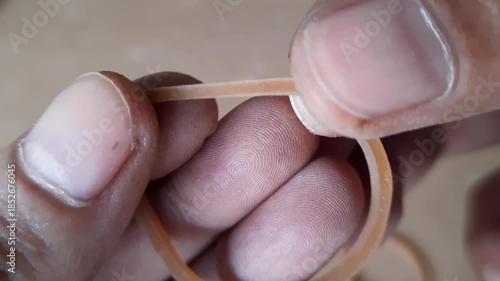 Two human hands pulling and stretching a single brown elastic rubber band shown in a detailed macro shot