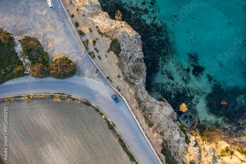 Aerial view of a car driving along a coastal road, where golden cliffs meet the turquoise sea, Popeye Village, Il-Mellieha, Malta.
