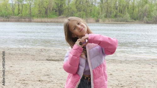 Joyful girl forms a heart with her hands on a sandy riverbank, smiling brightly in a pink jacket. A heartwarming scene of childhood happiness and love in nature.