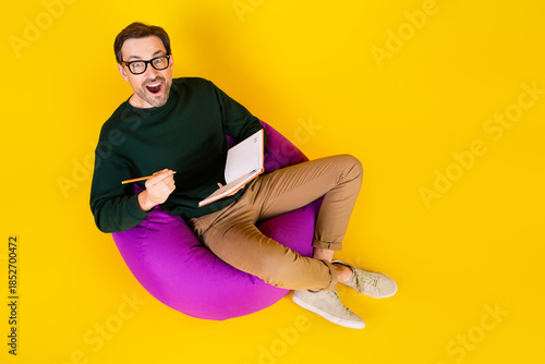 Photos Young man in green sweater sits on a purple beanbag writes in a notebook smiling