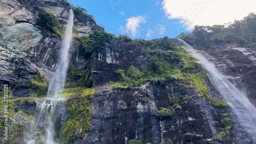 Looking up at towering twin waterfalls in Milford Sound, Fiordland National Park, New Zealand. Misty cliffs, cascading water, and dramatic fjord landscapes create a breathtaking scene.