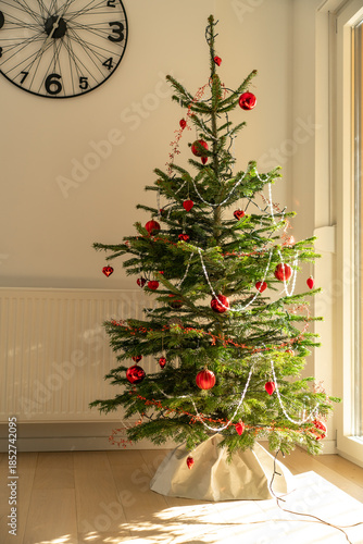 A simple natural Christmas tree decorated with red ornaments and light garlands standing in a bright modern interior. Cozy holiday atmosphere