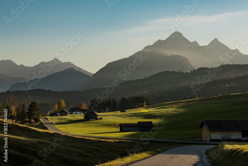 View of golden light illuminating the tranquil green meadows and scattered houses nested beneath the majestic, hazy mountains, Krun, Bavaria, Germany.