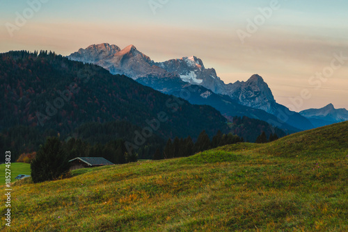 View of a rustic cabin nestled in a vibrant green meadow, backed by dark, dense forests and snow-dusted mountains under a pastel sky, Krun, Germany.