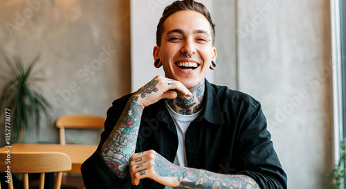 Smiling young man with tattoos, wearing a black shirt, sitting at a wooden table in a cozy cafe, showcasing a joyful expression and relaxed atmosphere