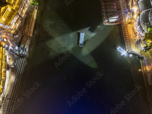 Aerial view of illuminated buildings casting golden light on the dark water, with a boat creating a focal point, Phoenix Ancient Town, Fenghuang County, Hunan, China.
