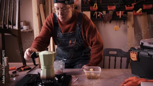 Female carpenter eating lunch and drinking fresh coffee at her workbench in small woodworking workshop