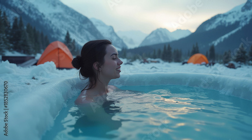 Woman soaking in a warm hot spring, snowy mountains and orange tents in the background, serene outdoor winter camping concept. Ice bath pod concept