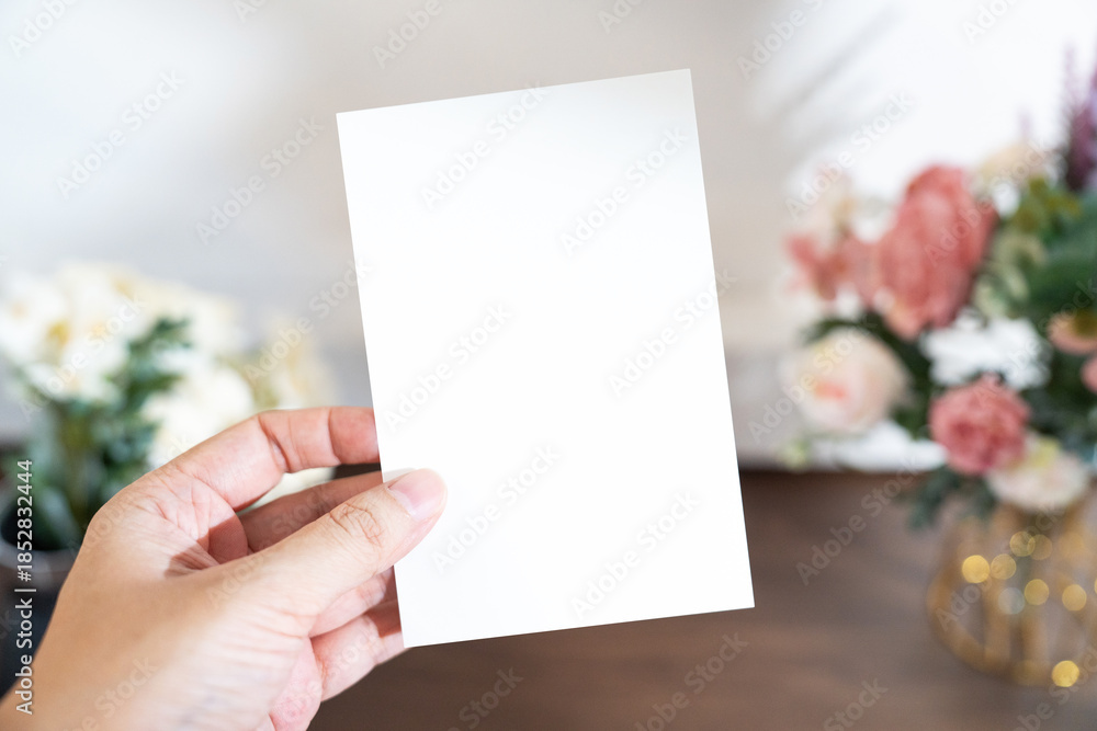 Obraz premium Mockup of a blank white card held by hand with soft blurred flowers, perfect for wedding or Valentine’s greeting design, with clipping path.
