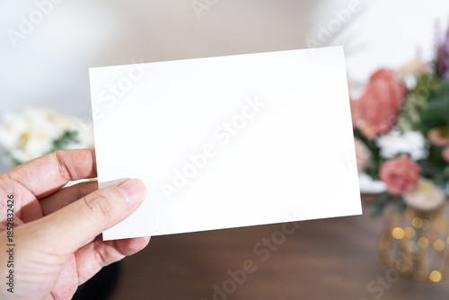 Mockup of a blank white card held by hand with soft blurred flowers, perfect for wedding or Valentine’s greeting design, with clipping path.