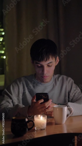 A young man sitting at a table in his cozy home during the evening, scrolling through his smartphone feed. Warm, soft lighting creates a relaxed and intimate atmosphere. Ideal for concepts of social m