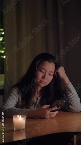 A young woman sitting at a table in her cozy home during the evening, scrolling through her smartphone feed. Warm, soft lighting creates a relaxed and intimate atmosphere. Ideal for concepts of social