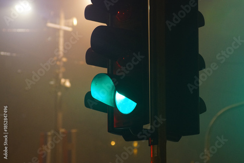 A green traffic signal glowing through dense urban fog during nighttime. Illustrates reduced visibility conditions, road safety management, smart city infrastructure, and climate-affected mobility.