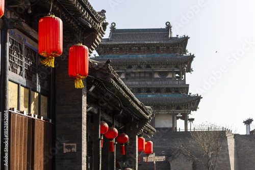 View of crimson lanterns casting a warm glow against the dark, textured walls of ancient buildings, with a majestic tower in the background, Pingyao, Shanxi, China.