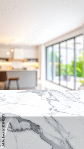 White marble countertop in a modern kitchen. Blurry background shows a bright interior, with kitchen cabinets, windows, and greenery