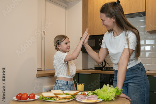 Mother and daughter are preparing sandwiches together in the kitchen, sharing a happy high five, symbolizing teamwork, achievement and bonding during healthy meal preparation