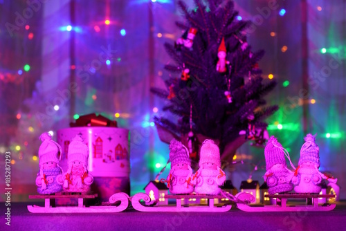 Toy angels and sledges on the background of a Christmas tree and a glowing garland. Purple color.
