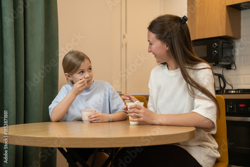 Mother and daughter sharing yogurt at the kitchen table, enjoying a casual, wholesome snack and warm conversation during a moment of family bonding and everyday togetherness