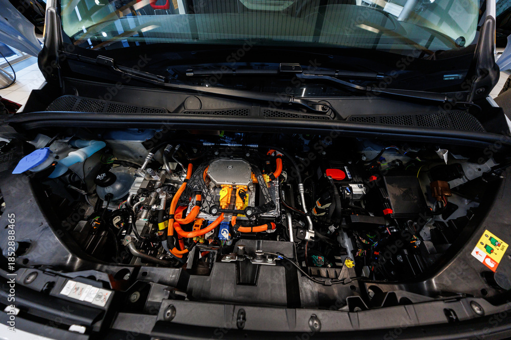 Obraz premium Open hood view shows orange high voltage cables, sealed inverter unit, coolant reservoirs, and a 12V battery indoors under showroom lighting during daytime.