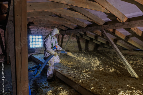 Worker in protective suit uses blue hose to spread loose fill insulation in a timber framed attic with exposed rafters and a glass block window under harsh side light.