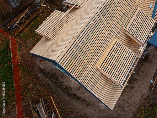 Tableau sur toile Aerial view shows a steep gable roof under construction with battens, dark underlayment, framed dormer openings, fallen leaves, ladders, stacked lumber, and a safety fence