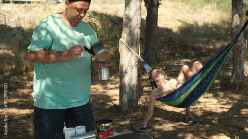 Father and son relax outdoors. Adult man boils water on a compact gas stove and pours it into mugs, teenage son swings and plays in a hanging hammock. unity with nature. Dream vacation
