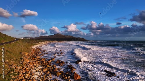 Aerial view of the rugged coastline where the wild, foamy waves crash against the rocky shore under a dynamic sky, Cape Town, Western Cape, South Africa.