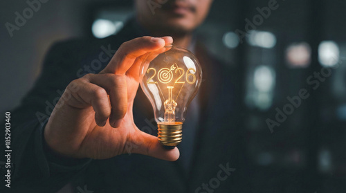 Businessman hand holds a glowing lightbulb with 2026 text and target icon. Concept of future business goals, innovation, and strategic planning