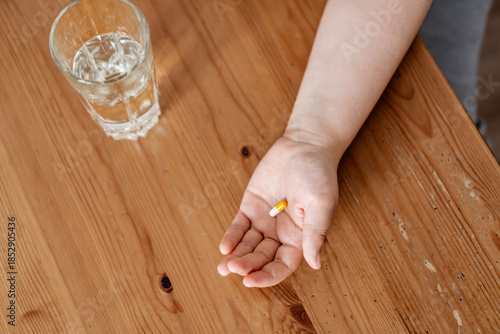 Kid holding capsule in the palm of his hand over tabletop with glass of water on the table with copy space. Concept of home treatment.