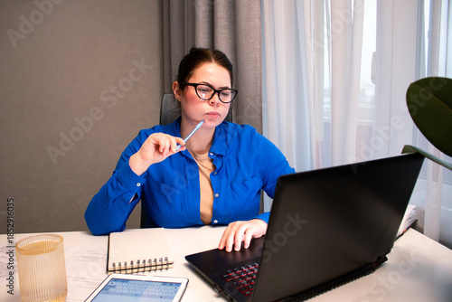 Concentrated woman with glasses types on laptop at home desk. Natural light from window highlights her productive remote work session. Focused young woman working remotely on laptop at home office