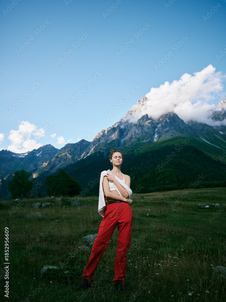 Naklejka premium Mountain landscape, outdoor fashion on a model in a grassy field with red pants and white top, dramatic sky and distant peaks, nature and style in color