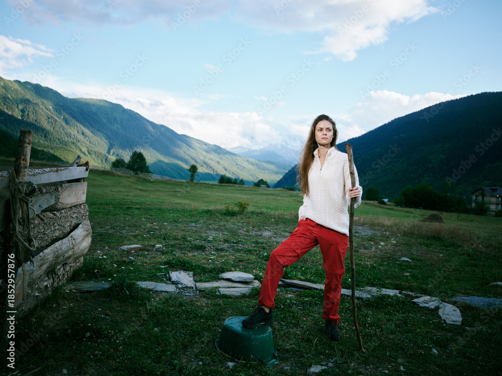 Naklejka premium Woman standing with a walking stick in a wide alpine meadow, mountains in the background, clear sky, fence on the left, rugged terrain, casual outdoor gear, calm countryside scene, exploring nature