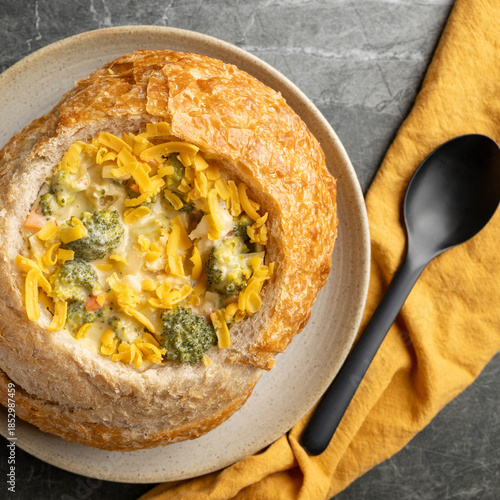 Broccoli cheddar soup in a sourdough bread bowl on marble surface with yellow cloth and black spoon.