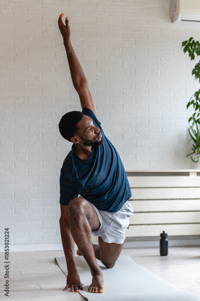 Naklejka premium Fit man stretching on a yoga mat at home, performing a side bend exercise after workout in a bright modern living room with plants, focused on flexibility and healthy lifestyle