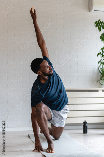 Fit man stretching on a yoga mat at home, performing a side bend exercise after workout in a bright modern living room with plants, focused on flexibility and healthy lifestyle