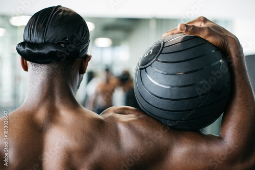 Athletic man holding medicine ball from behind in gym. Rear view of an athletic man holding a medicine ball during functional strength training in a modern gym.