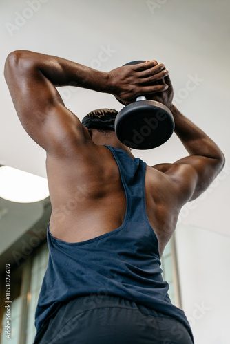Muscular man lifting weights. Muscular athlete training triceps with overhead dumbbell exercise in gym.